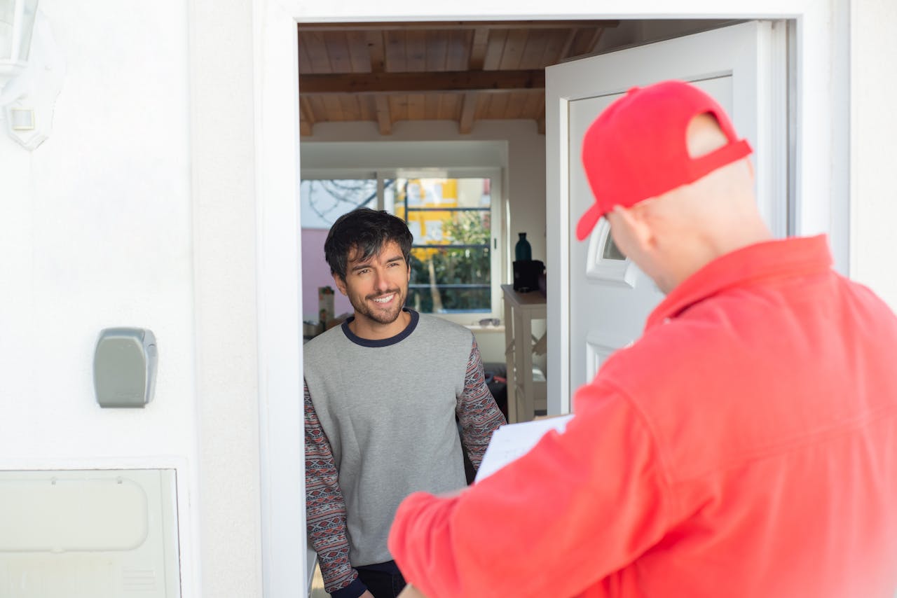 A delivery man in red uniform delivers a package to a smiling man at home.